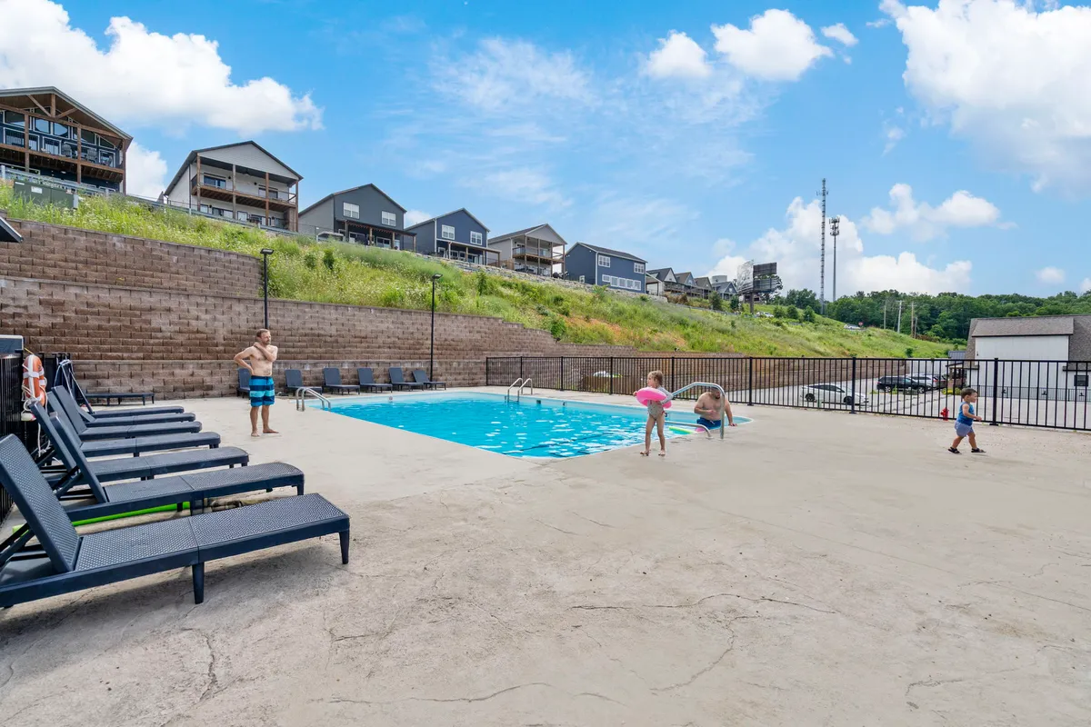 An active community pool scene with children splashing in the water, lounge chairs arranged for sunbathing, and scenic hills in the background. The mood is joyful and carefree, suggesting shared recreation and family-friendly fun.