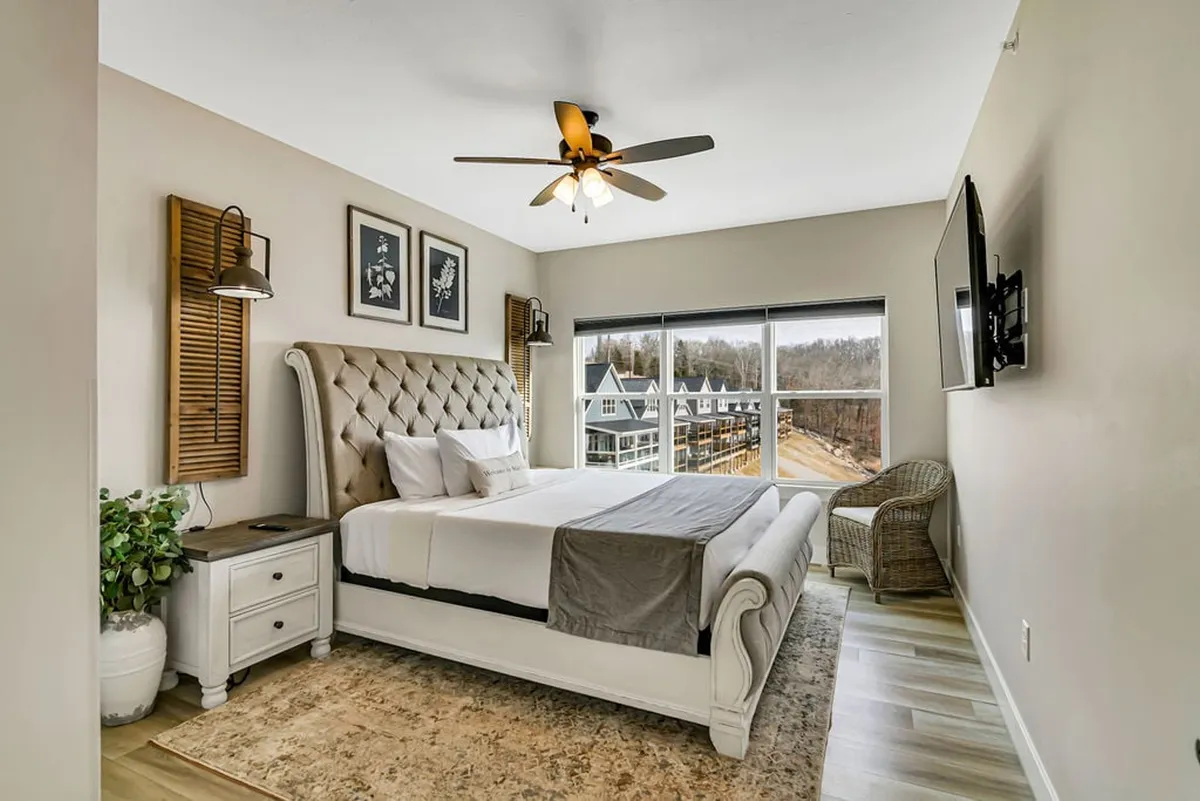 A serene bedroom with a plush tufted bed, large window showcasing a scenic view, decorative wall art, a modern ceiling fan, and a cozy rattan chair. The natural light and soft decor create an inviting, restful atmosphere.