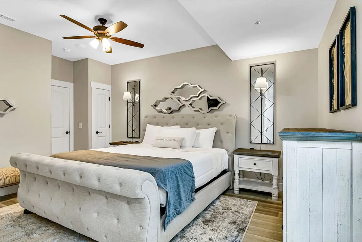 A tranquil bedroom highlighted by a comfortable bed, decorative mirrors, nightstands, and a ceiling fan. The space is bathed in natural light, perfect for curling up with a book or unwinding after a busy day.