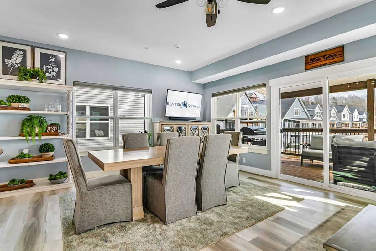 A comfortable, contemporary dining area with a prominent table, shelves adorned with plants, large windows offering lake views, and a visible TV screen. The scene invites thoughts of meals, games, and shared celebrations.