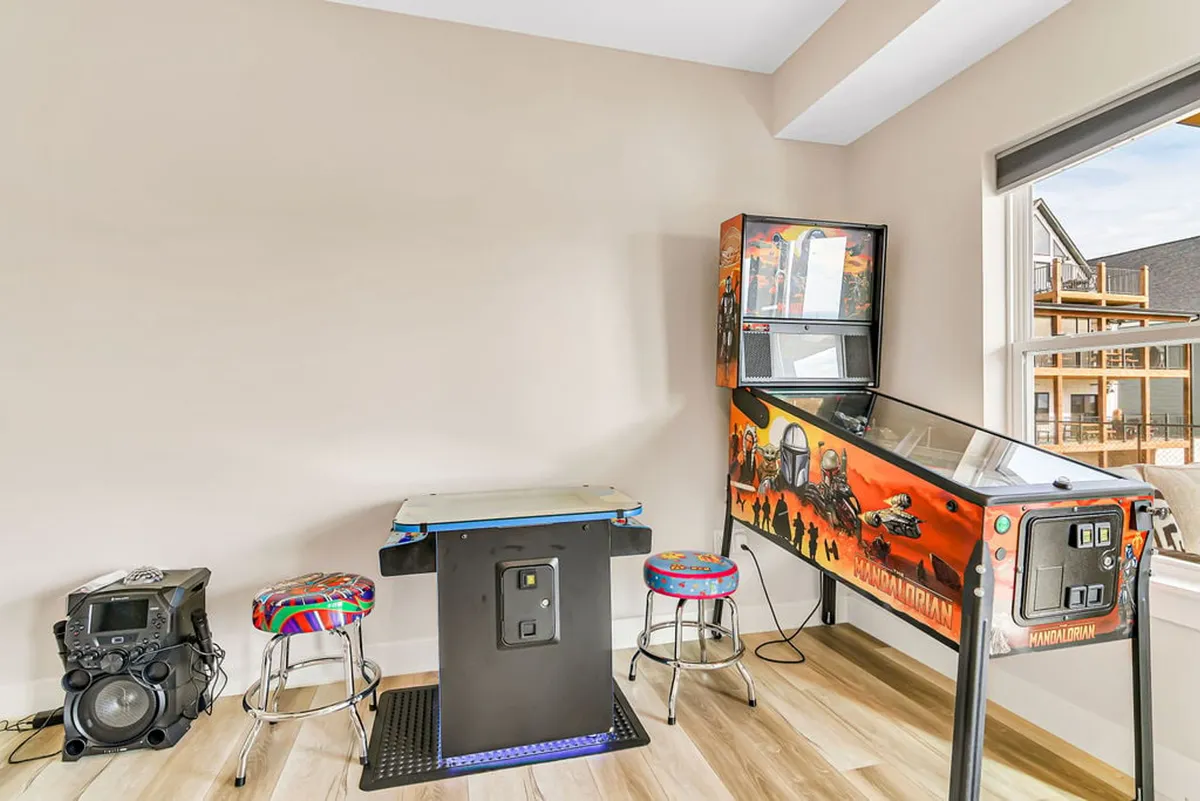 A vibrant interior game room featuring a retro arcade machine, a pinball machine, colorful stools, and a sleek sound system. The space feels lively and energetic, inviting guests to immerse themselves in entertainment and fun.