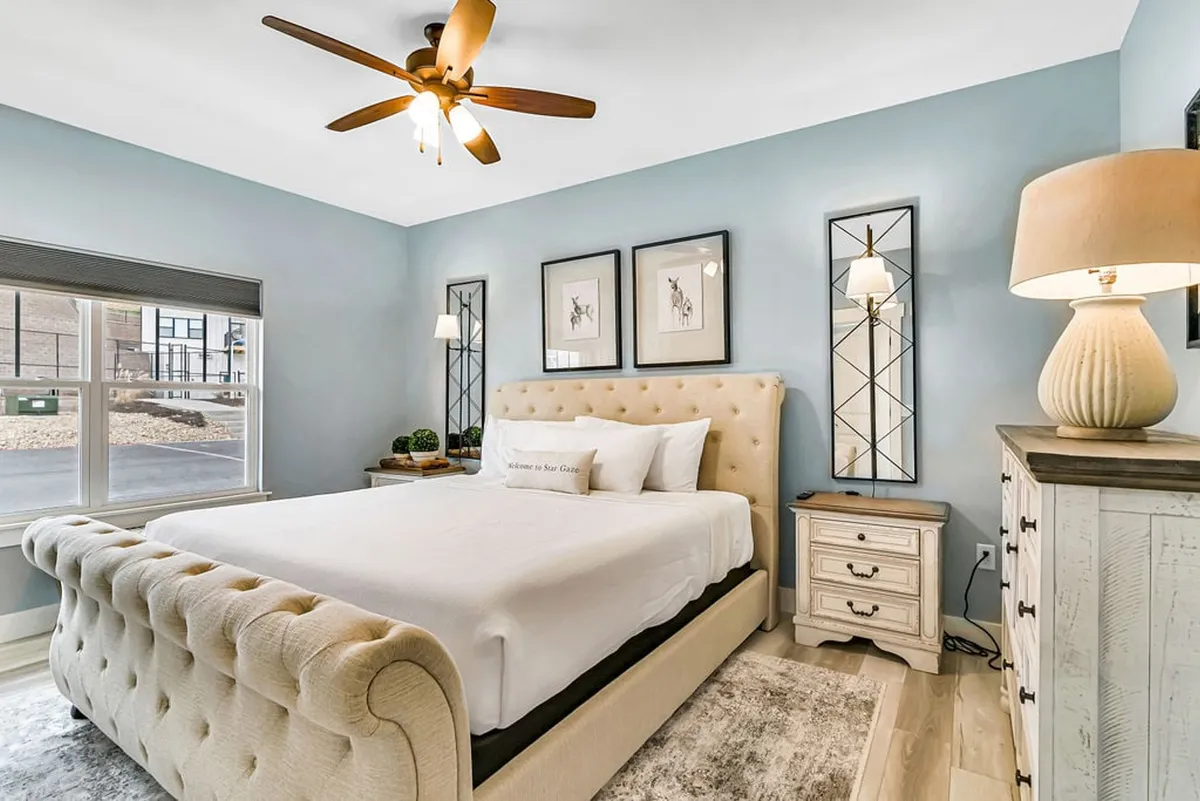 An elegant bedroom featuring a tufted bed, decorative wall art, bedside lamps, and a chest of drawers. Gentle light floods the room, creating a peaceful space ideal for unwinding after a day of activities.