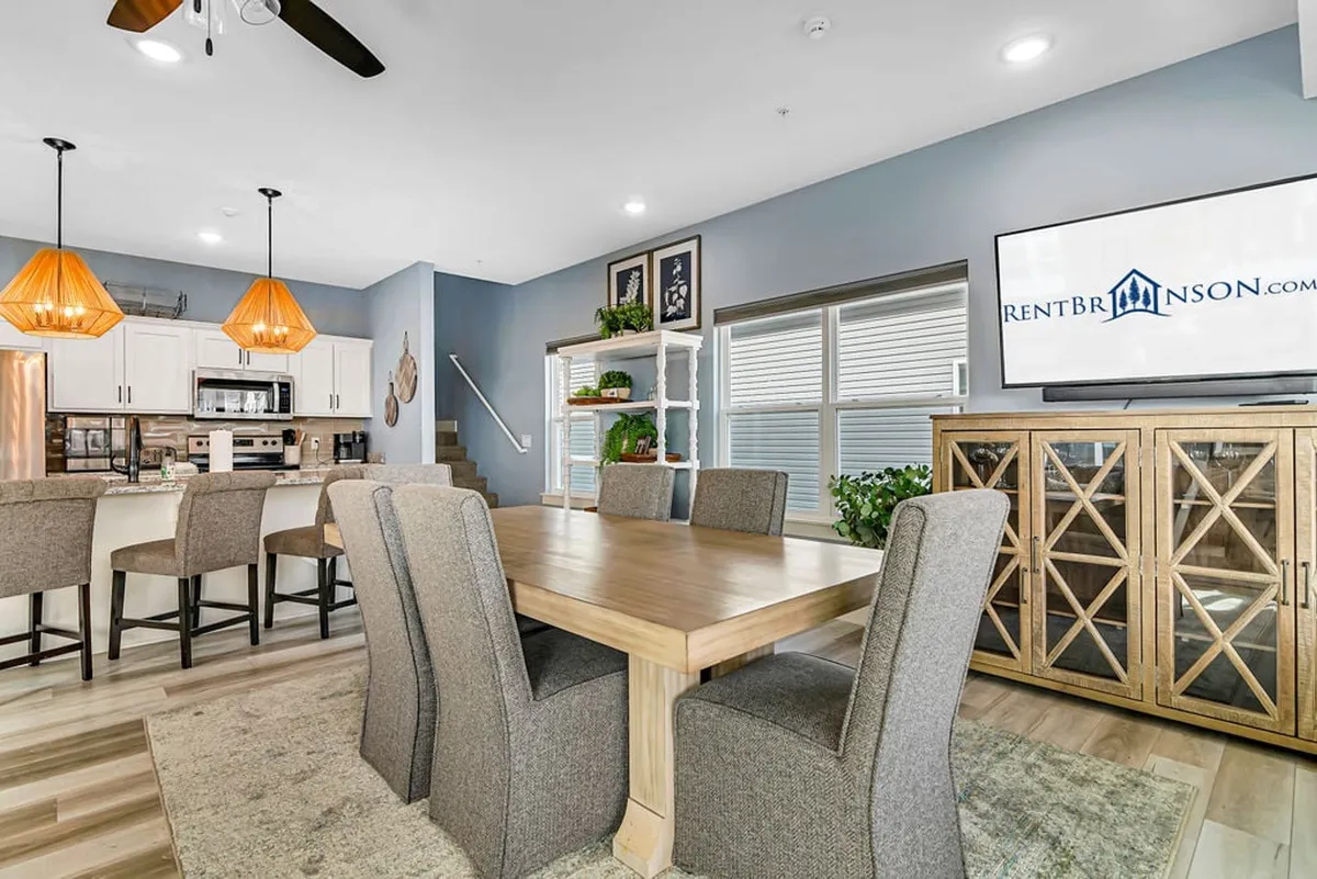 A warm, welcoming view of the great room that seamlessly connects the dining area, kitchen island, and living space. The shot shows plush seating facing a large television, a spacious dining table set before wide windows letting in natural light, and the open kitchen directly behind the dining space.