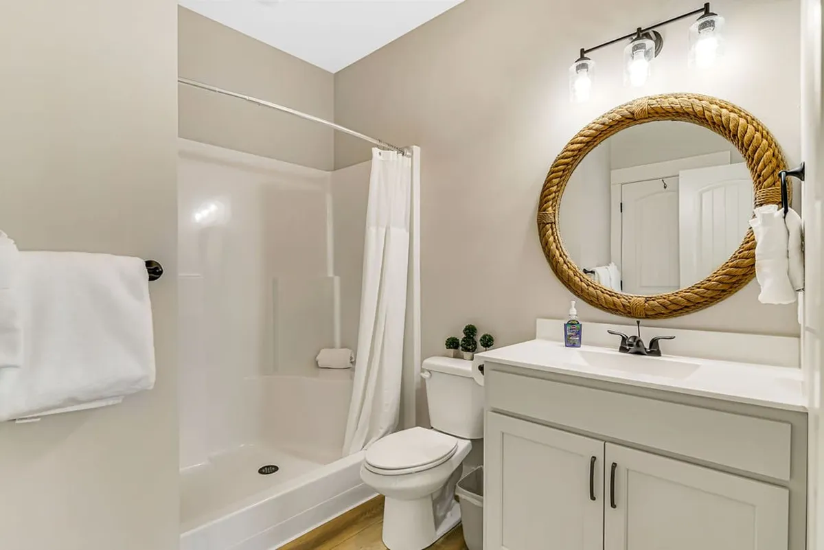 A stylish bathroom with a large round mirror framed in rope, a bright white vanity, and a shower area with a curtain. The space feels clean and spa-like, ideal for relaxation and self-care routines.