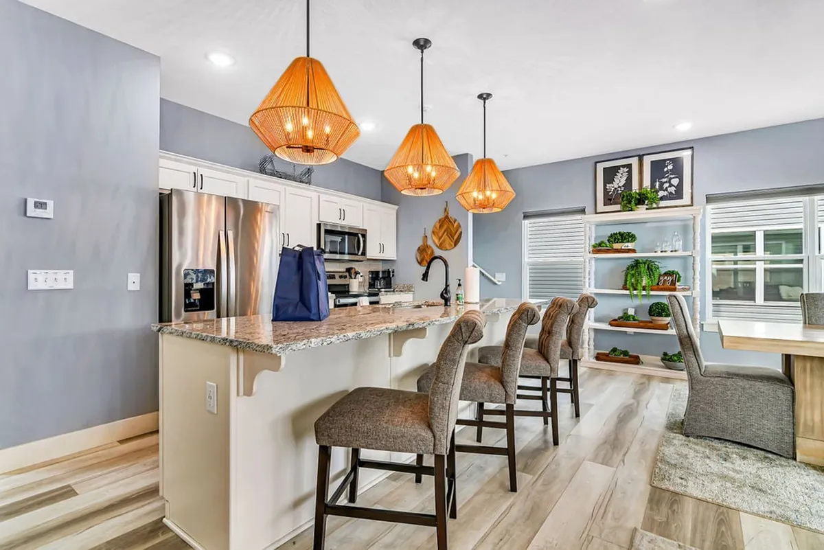 A welcoming scene highlighting the modern kitchen with a large island, hanging pendant lights, and a dining table nearby. The open shelving with plants and inviting layout create a homey environment ideal for family cooking and casual meals.