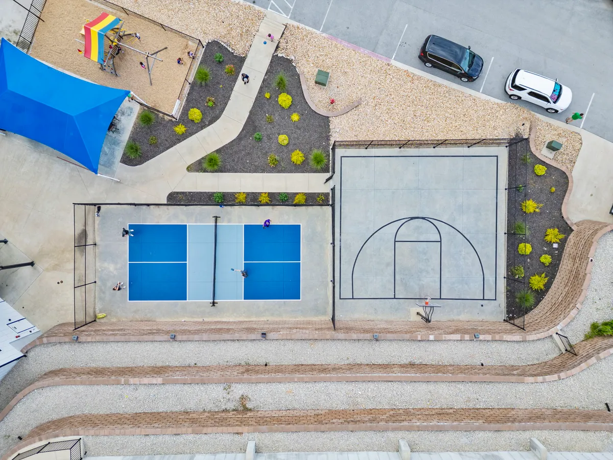 This exterior image showcases vibrant community amenities: tennis courts, a basketball court, and a playground area, all nestled together with a shade structure and landscaped gardens. Children and adults are seen enjoying various activities, highlighting the lively, family-friendly atmosphere.