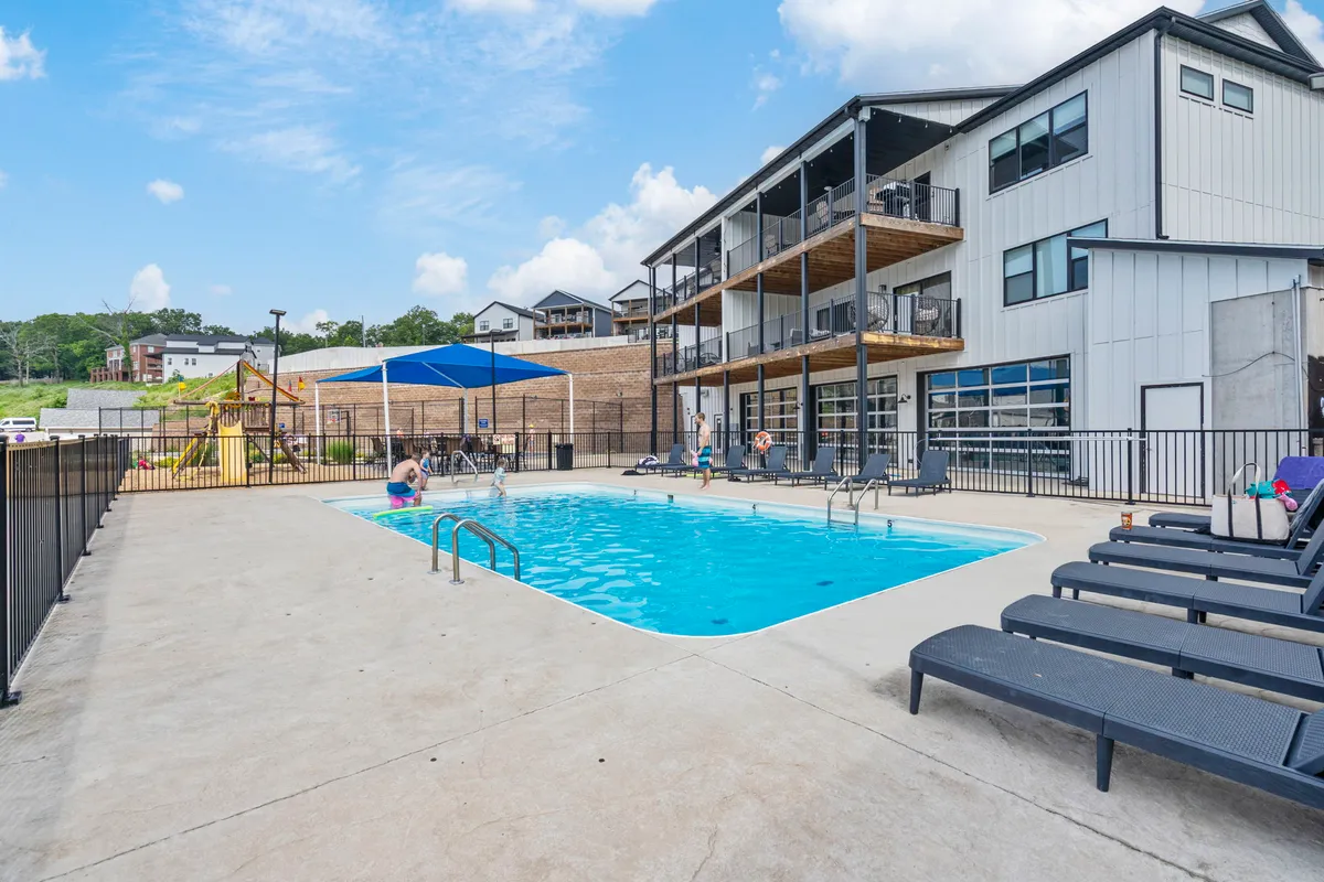 An inviting outdoor swimming pool scene with lounge chairs and a playground area beside the building's facade. The setting is cheerful and designed for fun, with families swimming, sunbathing, and children playing, suggesting easy access to both recreation and relaxation.
